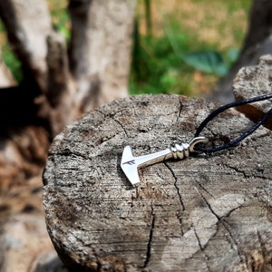 May include: A silver-colored pendant necklace featuring a hammer design, with a black cord. The hammer has a detailed handle and a stylized head with a small marking. The necklace is displayed on a weathered wooden surface.
