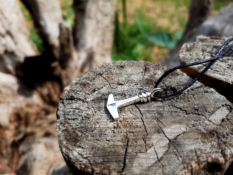 May include: A silver-colored pendant necklace featuring a hammer design, with a black cord. The hammer has a detailed handle and a stylized head with a small marking. The necklace is displayed on a weathered wooden surface.
