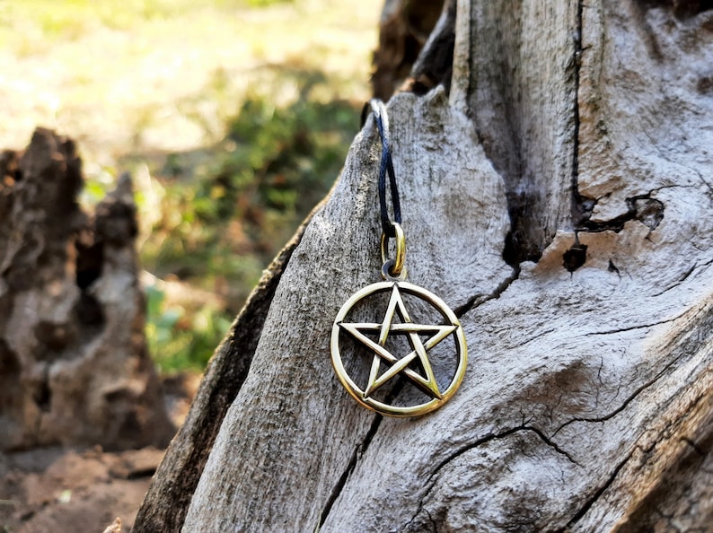 May include: A gold-colored pentagram pendant hangs from a black cord against a rustic wooden background.
