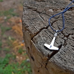 May include: A silver-colored pendant in the shape of Thor's hammer, with a blue cord. The hammer has a stylized design and a blue rune. The pendant rests on a weathered, textured wooden surface.