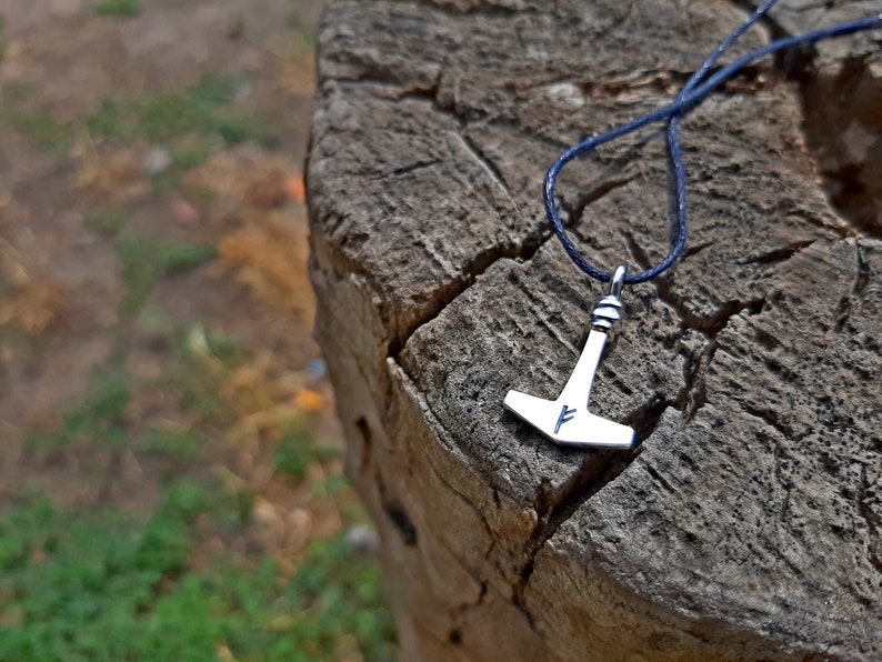 May include: A silver-colored pendant in the shape of Thor's hammer, with a blue cord. The hammer has a stylized design and a blue rune. The pendant rests on a weathered, textured wooden surface.