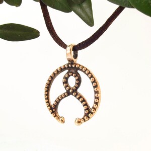 May include: A bronze pendant with a circular design featuring a stylized figure with a beaded border. The pendant is hanging from a brown cord.