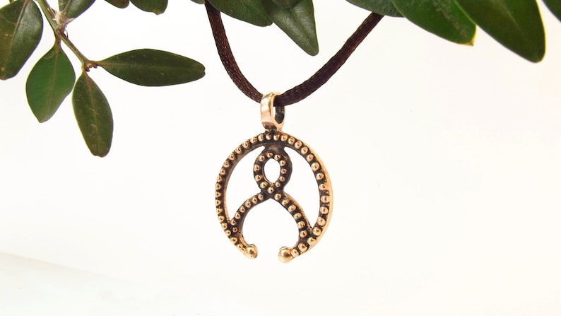 May include: A bronze pendant with a circular design featuring a stylized figure with a beaded border. The pendant is hanging from a brown cord.