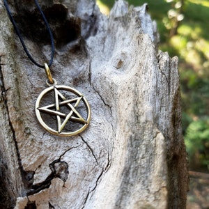 May include: A gold-colored pentagram pendant on a black cord necklace. The pendant is a five-pointed star with a circle around it. The necklace is lying on a piece of weathered wood.