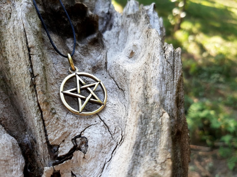 May include: A gold-colored pentagram pendant on a black cord necklace. The pendant is a five-pointed star with a circle around it. The necklace is lying on a piece of weathered wood.