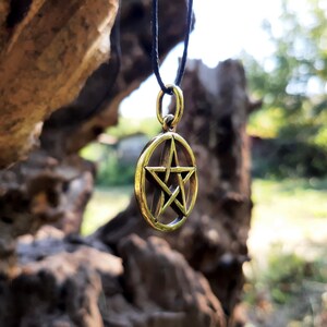 May include: A gold-colored pendant necklace with a pentagram charm. The charm is a five-pointed star inside a circle. The necklace is hanging from a black cord.