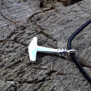 Op de afbeelding: Een zilveren hamer hanger aan een zwart leren koord. De hanger is een eenvoudig ontwerp met een afgeronde kop en een rechte steel.
