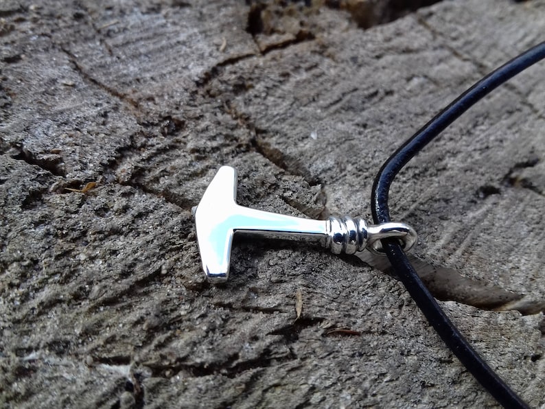 Op de afbeelding: Een zilveren hamer hanger aan een zwart leren koord. De hanger is een eenvoudig ontwerp met een afgeronde kop en een rechte steel.