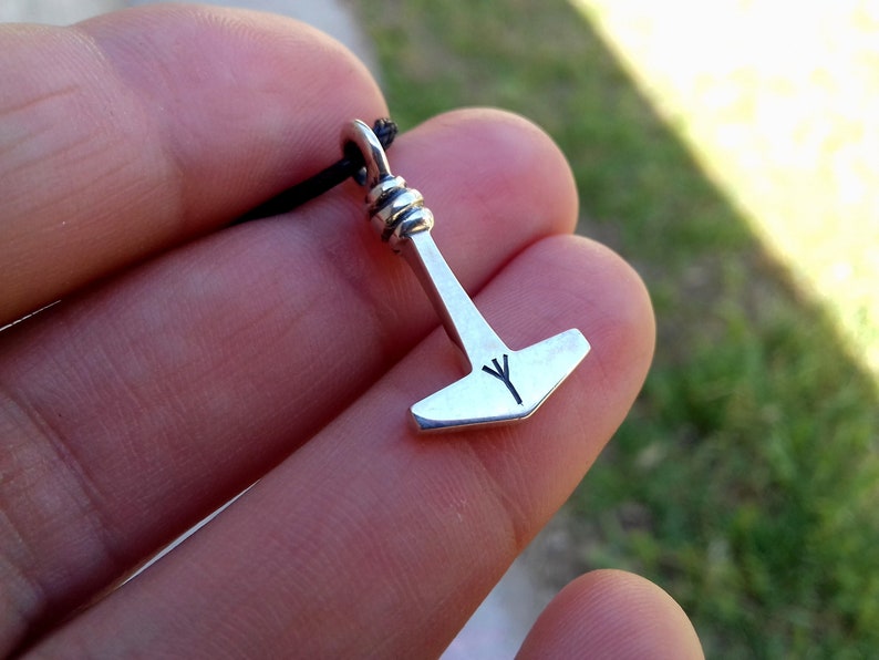 May include: A silver-colored pendant in the shape of Thor's hammer, with a black cord. The hammer has a stylized design and a rune symbol etched on the head. The pendant is held against a blurred green background.