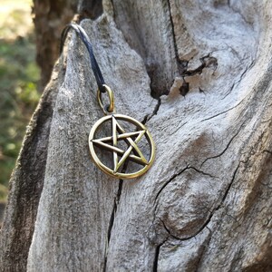 May include: A gold-colored pentagram pendant on a black cord, hanging from a piece of weathered wood.