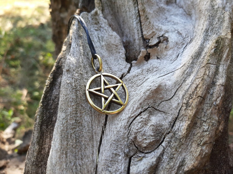 May include: A gold-colored pentagram pendant on a black cord, hanging from a piece of weathered wood.