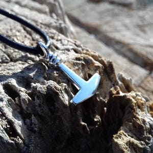 Op de afbeelding: Een zilverkleurige hamer hanger aan een zwart leren koord. De hanger is een eenvoudig ontwerp met een afgeronde kop en een rechte steel.