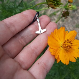 May include: A silver pendant in the shape of Thor's hammer, with a small rune, hangs from a black cord. The pendant is held near a bright orange flower with a yellow center, set against a green background.