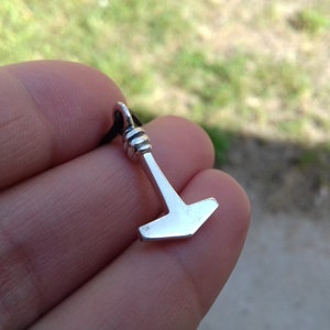 May include: A small, silver-colored pendant in the shape of a hammer, suspended from a black cord. The hammer has a detailed handle and a flat head. The pendant is held against a blurred green and gray background.