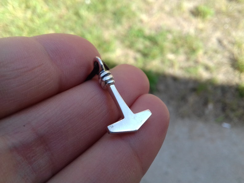 May include: A small, silver-colored pendant in the shape of a hammer, suspended from a black cord. The hammer has a detailed handle and a flat head. The pendant is held against a blurred green and gray background.