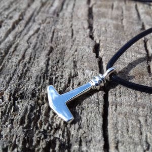 Op de afbeelding: Een zilveren hamer hanger aan een zwart lederen koordsnoer. De hanger is een eenvoudig ontwerp met een afgeronde kop en een rechte steel.