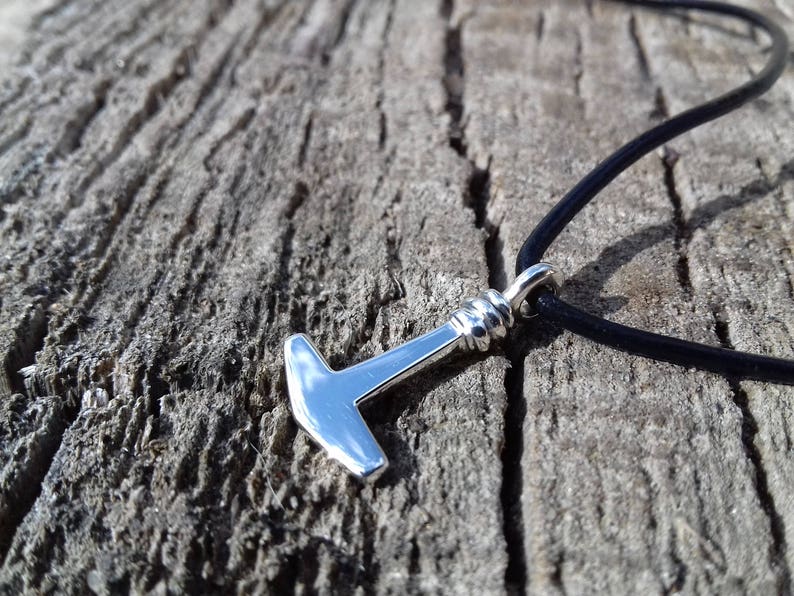 Op de afbeelding: Een zilveren hamer hanger aan een zwart lederen koordsnoer. De hanger is een eenvoudig ontwerp met een afgeronde kop en een rechte steel.