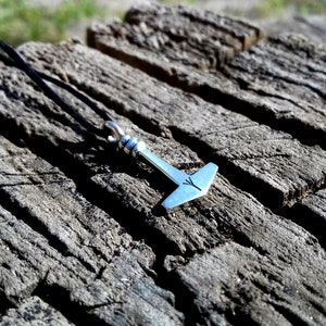 May include: A silver-colored pendant in the shape of Thor's hammer, with a black cord necklace. The hammer has a detailed design and a small rune symbol. The pendant rests on a weathered, gray wooden surface.