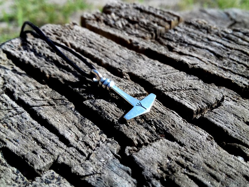 May include: A silver-colored pendant in the shape of Thor's hammer, with a black cord necklace. The hammer has a detailed design and a small rune symbol. The pendant rests on a weathered, gray wooden surface.