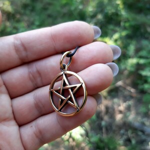 May include: A gold-toned pendant featuring a five-pointed star, also known as a pentagram, within a circle. The pendant is on a black cord.