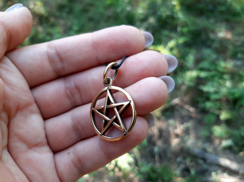 May include: A gold-toned pendant featuring a five-pointed star, also known as a pentagram, within a circle. The pendant is on a black cord.
