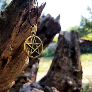 May include: A gold-colored pentagram pendant hangs from a black cord against a backdrop of a weathered tree trunk. The pendant is a five-pointed star within a circle.
