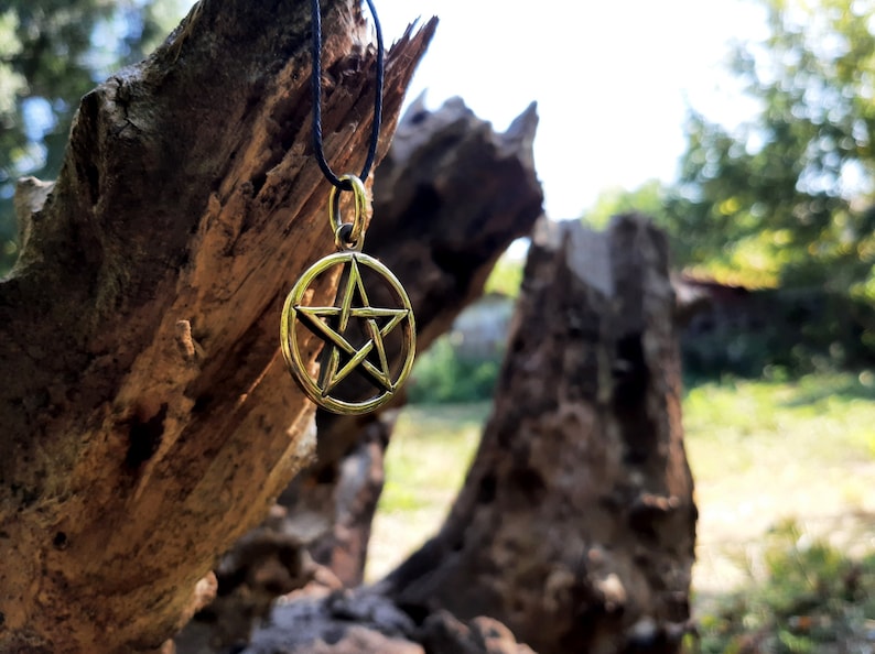 May include: A gold-colored pentagram pendant hangs from a black cord against a backdrop of a weathered tree trunk. The pendant is a five-pointed star within a circle.