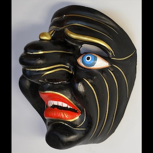 Puede incluir: Una máscara negra con detalles dorados y boca roja. La máscara presenta un rostro estilizado con un ojo azul y blanco. Probablemente para decoración o disfraces.