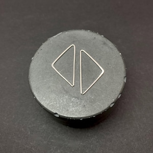 May include: A pair of minimalist silver triangle earrings displayed on a dark gray, circular surface. The earrings are simple geometric shapes, reflecting light. The background is a solid black, creating a contrast that highlights the jewelry.