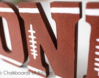 May include: Close-up of the letters "O" and "N" from the word "ONE" made of brown wood painted to look like a football. The letters have white stitching and white stripes around the edges.
