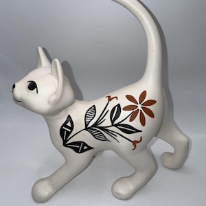 Puede incluir: Una figura de gato de cerámica blanca de pie con la cola arqueada hacia arriba. El cuerpo está decorado con un patrón floral negro y marrón de hojas y una flor. El gato tiene ojos pintados de negro y una expresión facial sencilla.