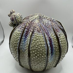 Puede incluir: Una escultura de cerámica redonda y texturizada que se asemeja a un erizo de mar. La superficie está cubierta de pequeñas protuberancias y presenta profundos surcos verticales esmaltados en tonos marrón oscuro, morado y azul, contrastando con el cuerpo pálido.