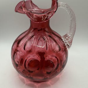 May include: A cranberry-colored glass pitcher with a ruffled rim and a clear, twisted glass handle. The body of the pitcher has a textured, bubble-like pattern. The pitcher is likely used for serving beverages.