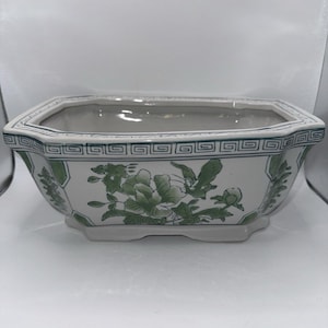 May include: A rectangular ceramic planter with a white base and green floral illustrations. The rim features a geometric Greek key pattern in green. The vessel has a wide opening, tapered sides, and a small footed base, showcasing a botanical design.