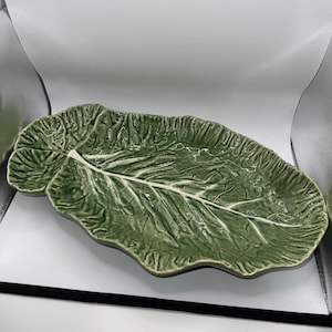 Peut inclure: Un plat de service en céramique verte brillante en forme de grande feuille de chou texturée. L'objet présente un motif de nervures en relief réaliste qui traverse le centre et longe les bords, imitant l'aspect naturel d'un légume.