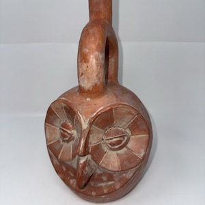 May include: A terracotta owl-shaped vessel with a handle. The owl's face is detailed with large, circular eyes and a beak. The vessel is a reddish-brown color with some lighter areas.
