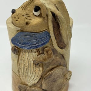 May include: A ceramic rabbit figurine with a blue scarf. The rabbit is tan with brown accents and has long ears. The figurine is cylindrical and has a textured surface.
