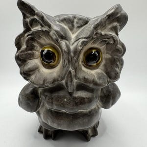 Puede incluir: Una figura de búho de cerámica con un acabado texturizado en gris y carbón moteado. El búho tiene ojos grandes y prominentes con pupilas brillantes de color marrón oscuro e iris ámbar, dentro de rasgos faciales profundamente esculpidos.