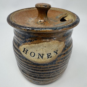 May include: A handcrafted ceramic honey pot with a lid and a small handle. The pot is brown with blue and black stripes and the word "HONEY" is printed on the front. The lid has a small opening for a honey dipper.
