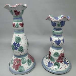 Puede incluir: Dos candelabros de cerámica con base blanca, adornados con corazones rojos, flores azules y rosas rosas. Los bordes están festoneados y pintados con detalles en azul y rosa. Los candelabros tienen un diseño caprichoso pintado a mano.