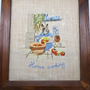 May include: A framed embroidery on beige fabric featuring a rabbit in a blue outfit preparing food in a kitchen. The scene includes a bowl of red produce, a brown pot, and blue kitchen items on a shelf. The text Home cooking is stitched in blue.