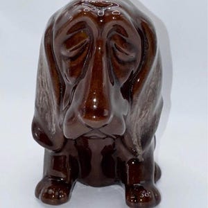 May include: A glossy, dark brown ceramic figurine of a basset hound dog. The dog is depicted in a seated position with long, drooping ears, a wrinkled forehead, and a downward-facing expression, featuring a smooth, reflective glazed finish.