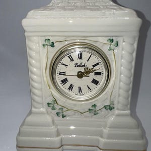 May include: A white ceramic mantel clock with a round clock face. The clock face has black Roman numerals and gold hands. The clock is decorated with green shamrocks and the word "Belleek".