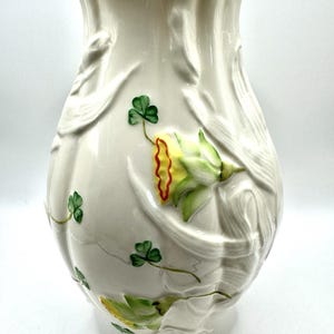 May include: A cream-colored ceramic vase with a scalloped rim and a relief-molded surface featuring raised, hand-painted green shamrocks and yellow daffodils with orange-red centers. The vase has a glossy finish and a bulbous, tapered shape.