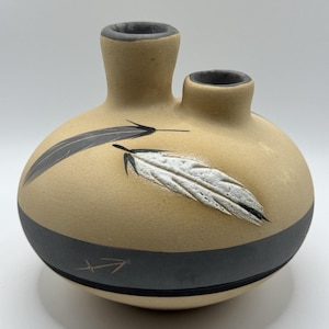 May include: A tan ceramic vase with two openings and a round body. The vase features a black band around the middle and is decorated with black and white feather designs. An arrow symbol is etched on the side.