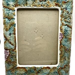 May include: A rectangular ceramic picture frame featuring a raised, three-dimensional relief pattern of grape leaves and clusters of grapes. The leaves are colored in shades of teal, green, and brown, while the grapes are a soft, muted purple.