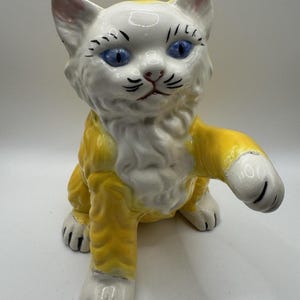 May include: A ceramic cat figurine featuring a white face and chest with yellow fur on its body and limbs. The cat has bright blue eyes, painted black eyelashes, whiskers, and a pink nose. One front paw is raised in a waving or reaching gesture.