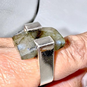 May include: A silver ring featuring a rectangular, faceted labradorite gemstone set horizontally. The stone displays grey and green tones with subtle iridescent flashes, held securely by two polished silver prongs on a wide, smooth silver band.