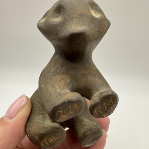 May include: A small, dark brown, matte-finished clay figurine of a stylized animal in a seated position. The base of the figurine features hand-etched text including the letters RM, the number 213, and the word ITALY inscribed on the bottom feet.