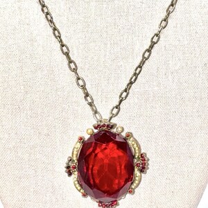 May include: A necklace featuring a large, oval-shaped, faceted red glass pendant set in a metallic frame. The pendant is accented with small, clear white stones and tiny red crystals along the edges. It hangs from a simple, dark-toned metal chain.